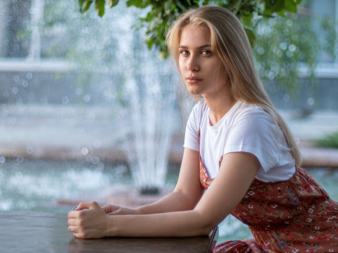 Slender charming young blonde in white T-shirt and multi-colored sundress poses in front of camera against  background of fountain in city park. Sunny day in city. falling splashes water lit by sun