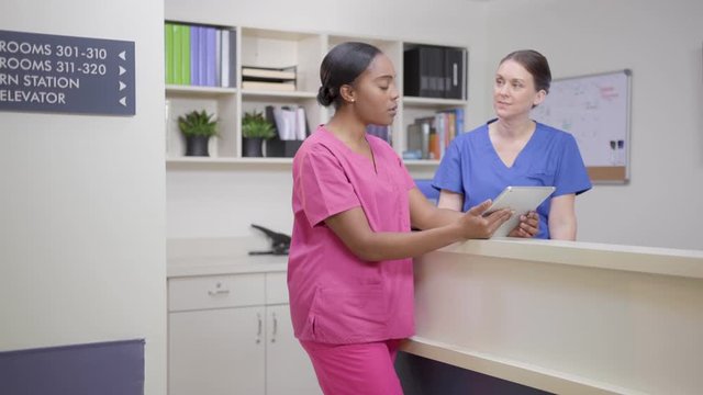 Two nurses at nurses station review patient chart on tablet, camera move left