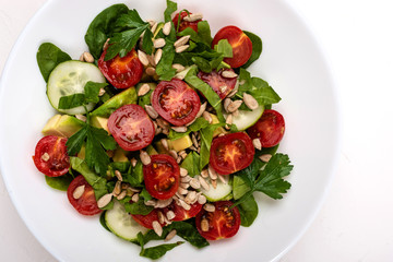 Salad with avocado, cucumbers, cherry tomatoes, spinach leaves, parsley and seeds on a white plate on a white background. Healthy vegetarian food.