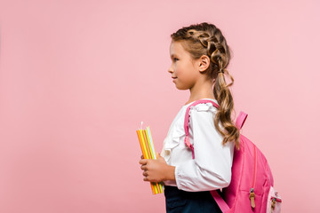 side view of happy kid holding books while standing with backpack isolated on pink