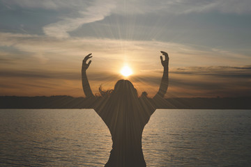 Silhouette of young woman which is looking at a sky in a sunset rays and is stretching her hands to...