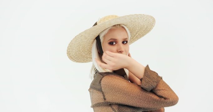 Pretty female model with blonde hair in straw hat and transparent clothing posing for camera isolated on white background
