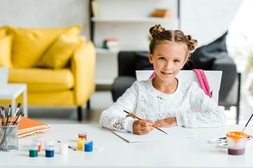 happy kid holding paintbrush near gouache jars and paper on table