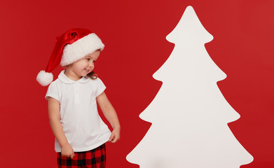 Happy little child girl in Santa hat at the Christmas time. Isolated red background. Empty place for an inscription in the form of a Christmas tree