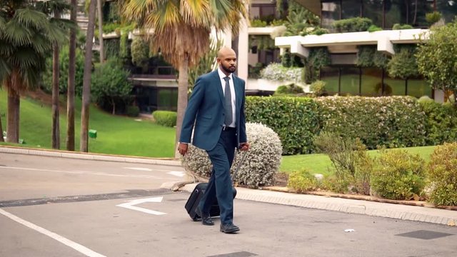 Indian businessman wearing blue suit walking with travel bag or suitcase near office or hotel. Travel business concept