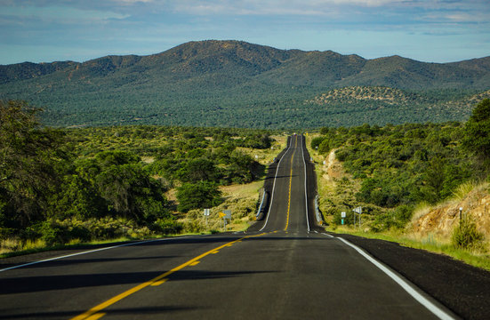 A straight expanse of highway on New Mexico state route 90, north of Lordsburg, south of Silver City through high country of mountains, juniper and pine trees under a summer sky