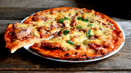 Homemade pizza with cheese, tomatoes, bacon and herbs in a white ceramic plate on a wooden table.