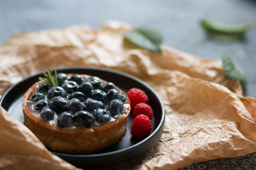 Delicious blueberry tartalets with vanilla cream and fresh cream with raspberry on a black and white plate on a grey background.