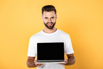 happy handsome man in white t-shirt showing laptop with blank screen isolated on yellow