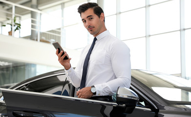 Young man with phone near car in modern dealership
