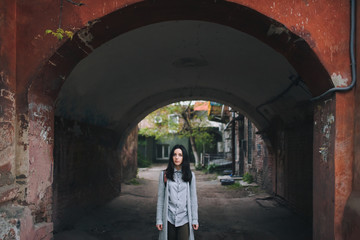 A young sad girl in a gray cardigan and a shirt stands in the arch of an old slum yard and looks at...