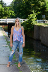 Beautiful young blonde woman poses outside in denim and print tank-top - near creek or canal