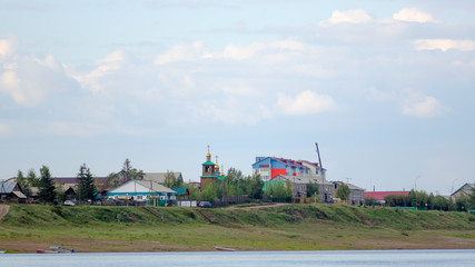Fototapeta premium Houses with silhouettes of cars on the road near the cliff of the Northern vilyu river go to the Orthodox Church with Golden domes among the houses in the wild North of Yakutia in the ulus Suntar.