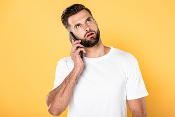 thoughtful handsome man in white t-shirt talking on smartphone isolated on yellow