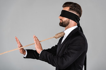 side view of blindfolded businessman in black suit with noose on neck showing stop gesture isolated on grey