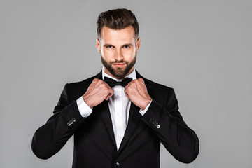elegant smiling man in black suit fixing bow tie isolated on grey