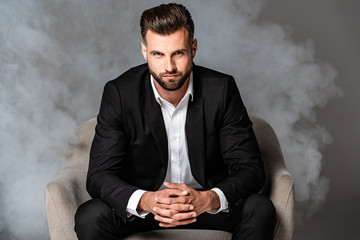handsome businessman in black suit sitting in armchair in smoke on grey background