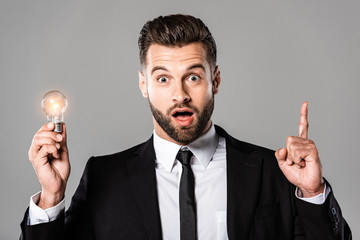 surprised businessman in black suit holding glowing light bulb and showing idea gesture isolated on...