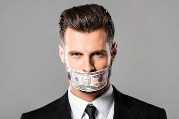 sly businessman in black suit with dollar banknote on mouth isolated on grey