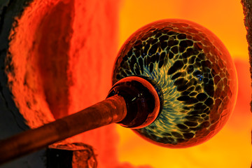 Glass blower working on a bubble of melted glass on a rod by heating it up in a kiln at a glass...