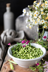 Young green peas in white bowl on wooden background. Pea flowers and daisy flowers on the table.