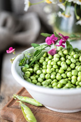 Young green peas in white bowl on wooden background. Pea flowers and daisy flowers on the table.
