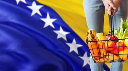 Woman is holding supermarket basket, Bosnia Herzegovina waving flag background. Economy concept for fresh fruits and vegetables.