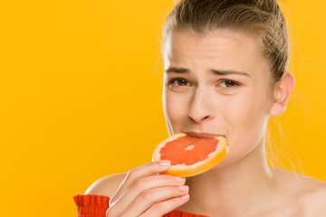 Young beautiful woman with towel on her head eat slice of grapefruit with sour gesture on yellow background