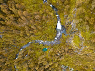 Obraz na płótnie Canvas A large waterfall in the back of the Altai Mountains near a steep cliff with yellow trees, house and green roof. Rest and loneliness while traveling to deserted places. Aerial view. Indian summer.