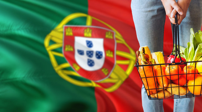 Woman is holding supermarket basket, Portugal waving flag background. Economy concept for fresh fruits and vegetables.