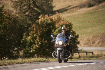 Bearded handsome motorcyclist in helmet, sunglasses and black leather clothing riding cruiser motorbike along empty asphalt road on bright sunny day.