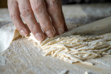 Senior woman cuts the dough on wooden board during homemade noodle or pasta production in her home kitchen. Closeup, selective focus