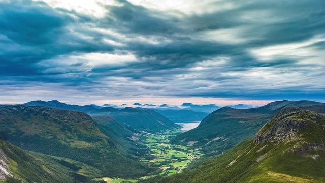 Dark stormy clouds passing over the narrow valley. Mountains towering on its' sides. Blue waters of fjord seen in the distance.