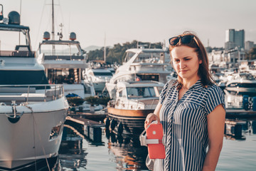 Cute girl in a striped dress makes selfie on a camera of her red smartphone with selfie stick on the background of sea and boats. Toned