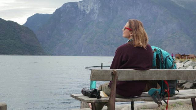 Young woman traveler sitting on the bench with a view to the fjord and resting, big backpack standing next to her.