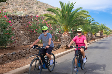 A senior couple man and woman enjoying freedom with electric bicycle. Happiness, fun and emotion outdoor in the street. Flowering plants and palm trees in background