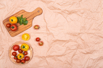 Fresh ripe small tomatoes in wooden plate and on olive wood cutting board on brown crumpled paper background. View from above.