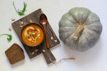 Roasted pumpkin and carrot soup with cream, black pepper and pumpkin seeds ,cutting board and fresh pumpkin slices,black bread on white concrete background.