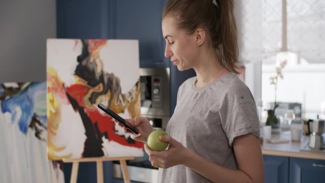 Young female painter eating green apple and scrolling through social media on smartphone while standing in home art studio