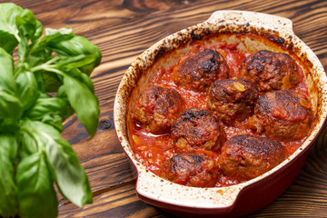 Meatballs in tomato sauce in a baking bowl, wooden table. View from above. basil in the foreground in defocus. baked meatballs/