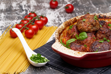 meatballs with tomato sauce and decorated with basil leaf, served in white and red pan on grey background. next to the pan near fresh tomatoes, spaghetti. Closeup.