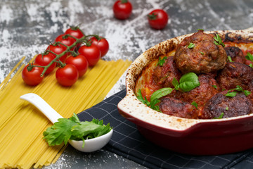 meatballs with tomato sauce and decorated with basil leaf, served in white and red pan on grey background. next to the pan near fresh tomatoes, spaghetti. Closeup.