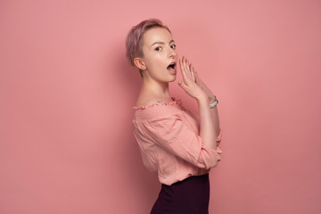 Fototapeta premium A girl with short hair is standing in profile on a pink background and looking at the camera with an embarrassed cover for mouth