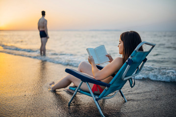 girl sitting on a beach chair reading a book on the shore, there is a boy away from her