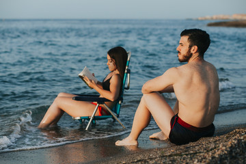 couple enjoying a beach day - she reads a book while he looks at the horizon