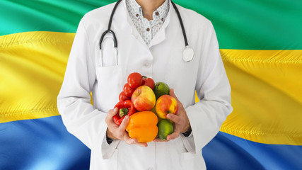 Doctor is holding fruits and vegetables in hands with Gabon flag background. National healthcare...