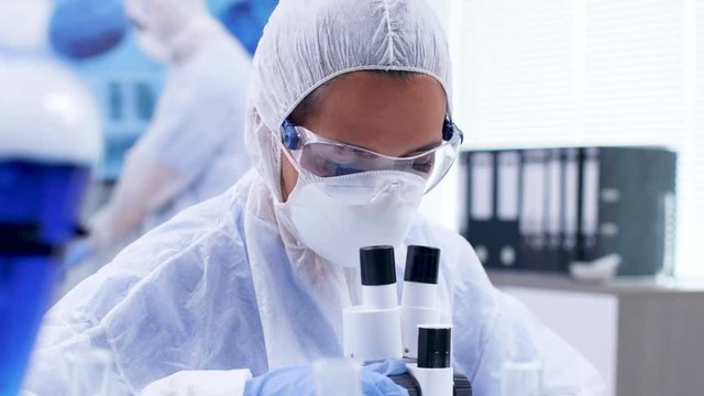 Close up footage of female scientist looking through moden microscope in research laboratory. Scientist in coverall protection equipment.