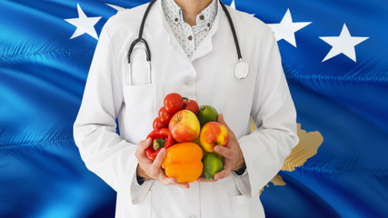 Doctor is holding fruits and vegetables in hands with Kosovo flag background. National healthcare concept, medical theme.