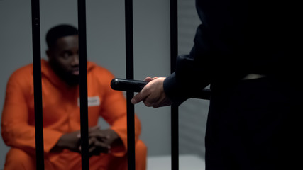 Afro-american prisoner in cell looking at jail guard with baton, harassment