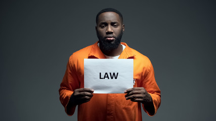 Black imprisoned man holding law sign in cell, looking to camera, human rights